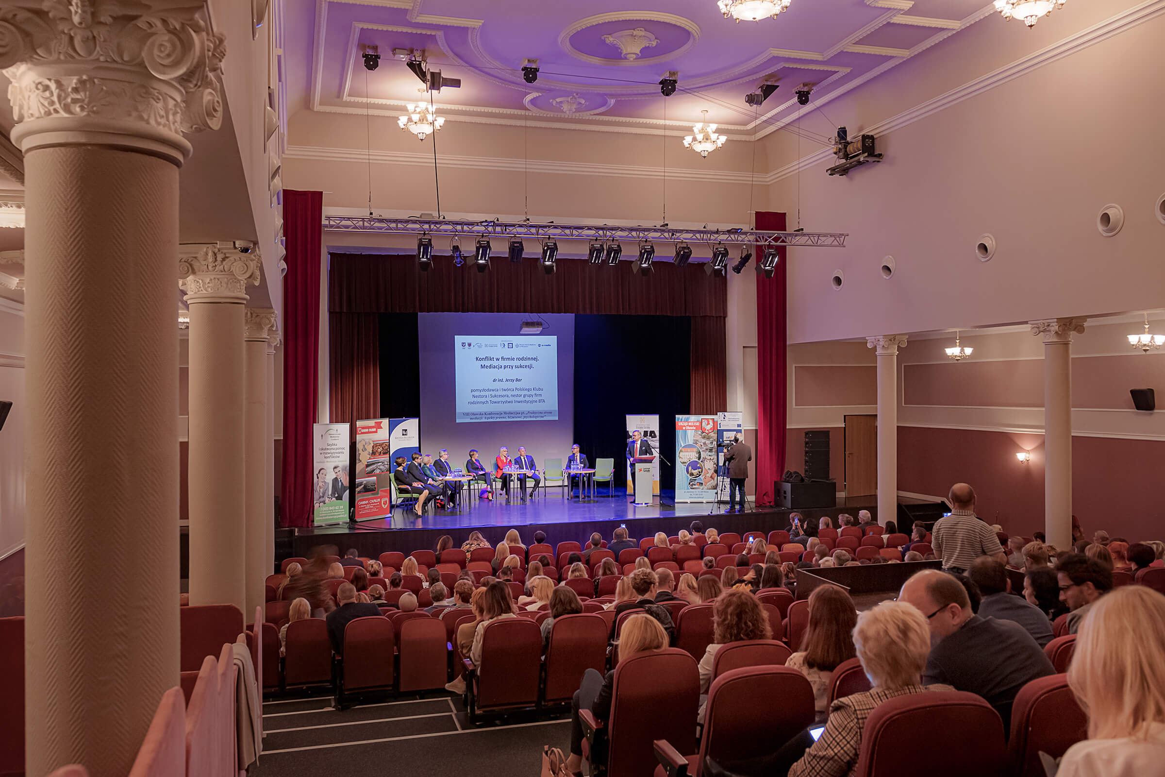 Grupa ludzi siedzi w sali widowiskowej. Na scenie przy stolikach siedzi osiem osób, przy mównicy jedna osoba, kamerzysta nagrywa wydarzenie. Tłem sceny są rolapy reklamowe i slajd na ekranie.