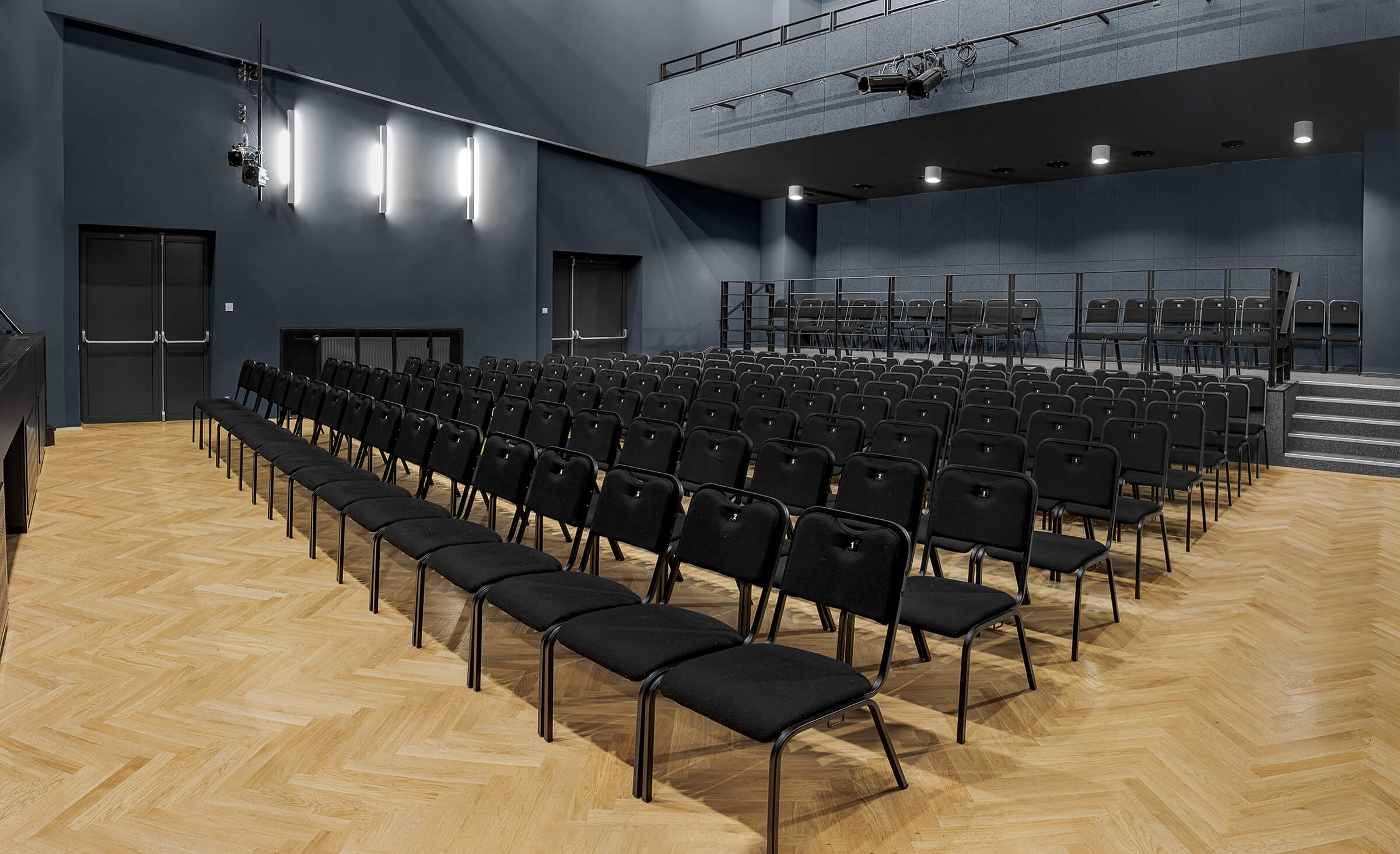 Sala widowiskowa z rzędami krzeseł, za nimi, po obu stronach są schody prowadzące na podwyższenie z krzesłami. W tle dwie pary drzwi.