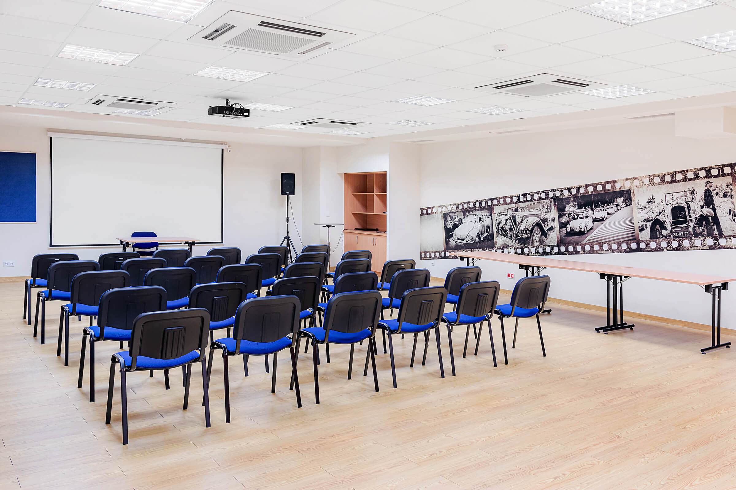 Sala konferencyjna. Rzędy krzeseł, przed którymi wisi ekran i stoi głośnik. Na ścianie fototapeta ze wzorem starych aut i szafa.