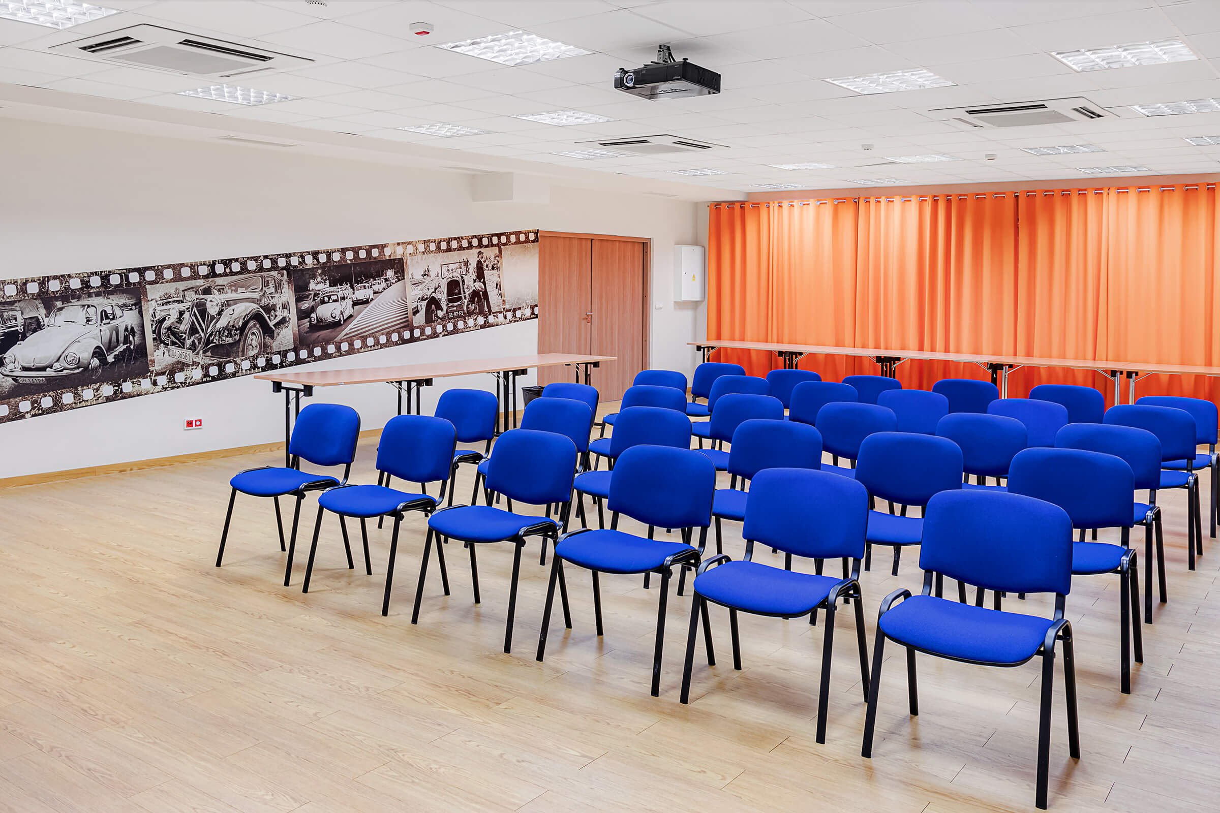 Sala konferencyjna, a w niej pięć rzędów krzeseł. Na ścianie fototapeta ze wzorem starych samochodów. Za krzesłami stoją stoły.
