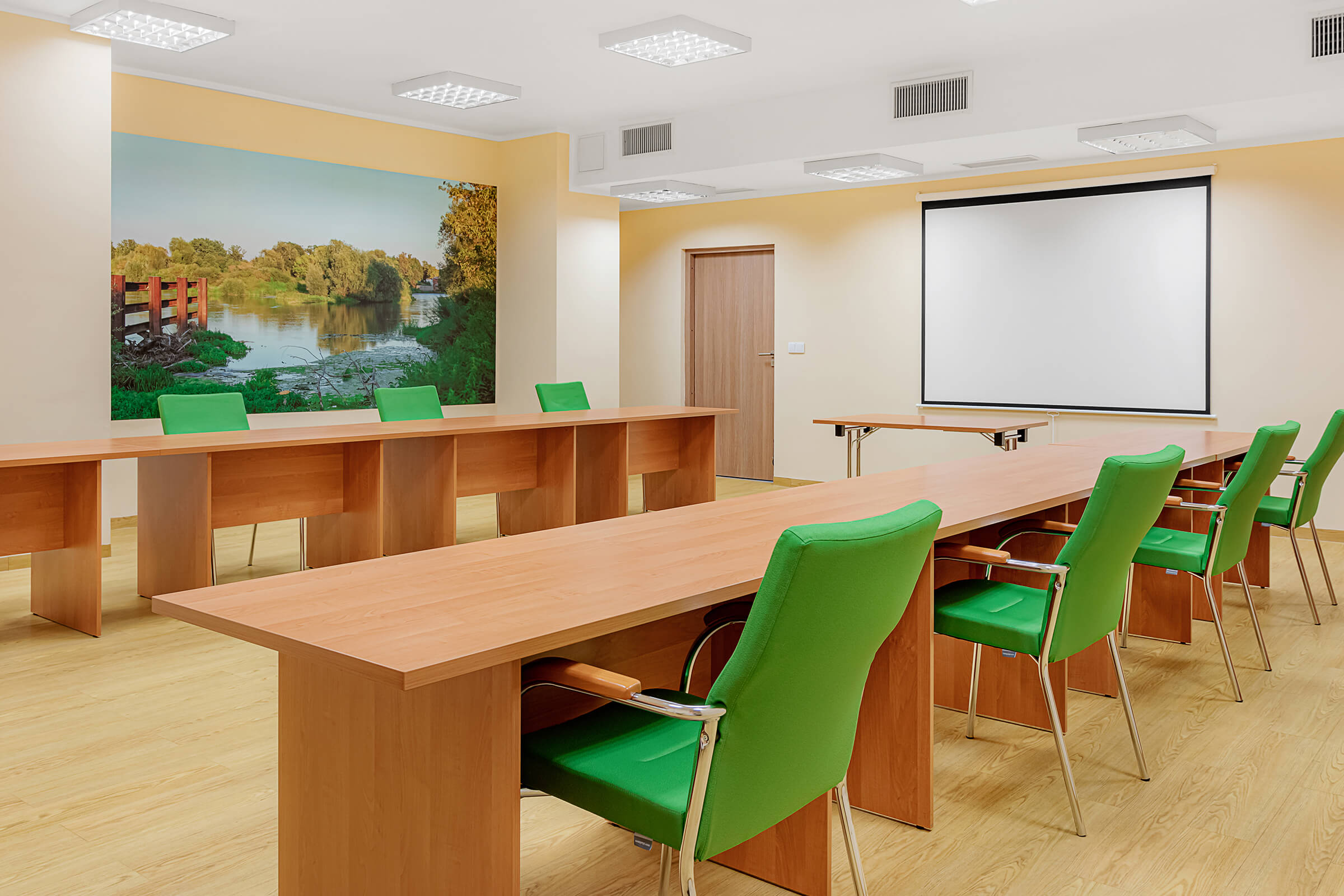 Sala konferencyjna. Na ścianie fototapeta z drzewami i rzeką. Stoły ustawione w rzędach na przeciwko siebie. Na ścianie ekran.