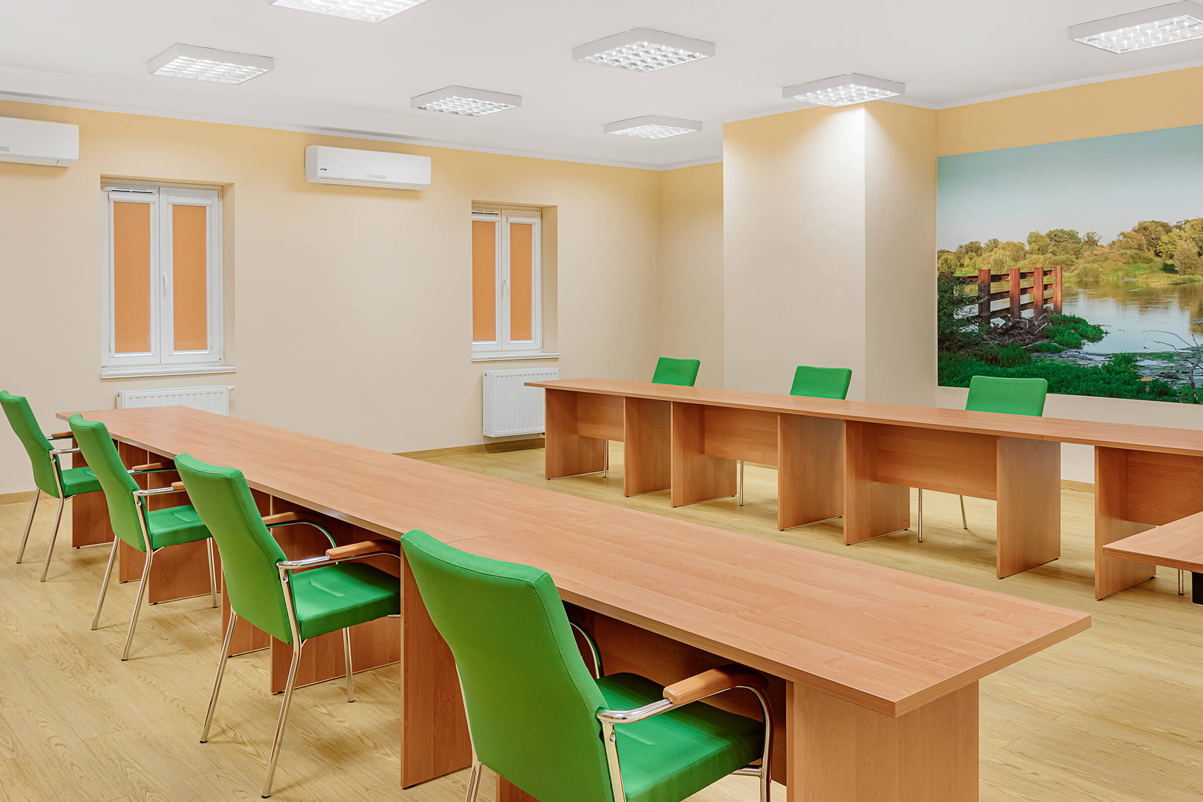 Sala konferencyjna. Na ścanie fototapeta z drzewami i rzeką. Stoły ustawione w rzędach na przeciwko siebie. Dwa okna z zasłoniętymi roletami.