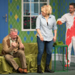 na pierwszym planie od lewej na sofie siedzi płaczący mężczyzna w garniturze, po środku stoi kobieta w dżinsach i koszuli obok telefonu stacjonarnego, po prawej mężczyzna ma okulary, ubrany w ubrudzony kombinezon z unosi ręce, w tle okno z widokiem na ogród